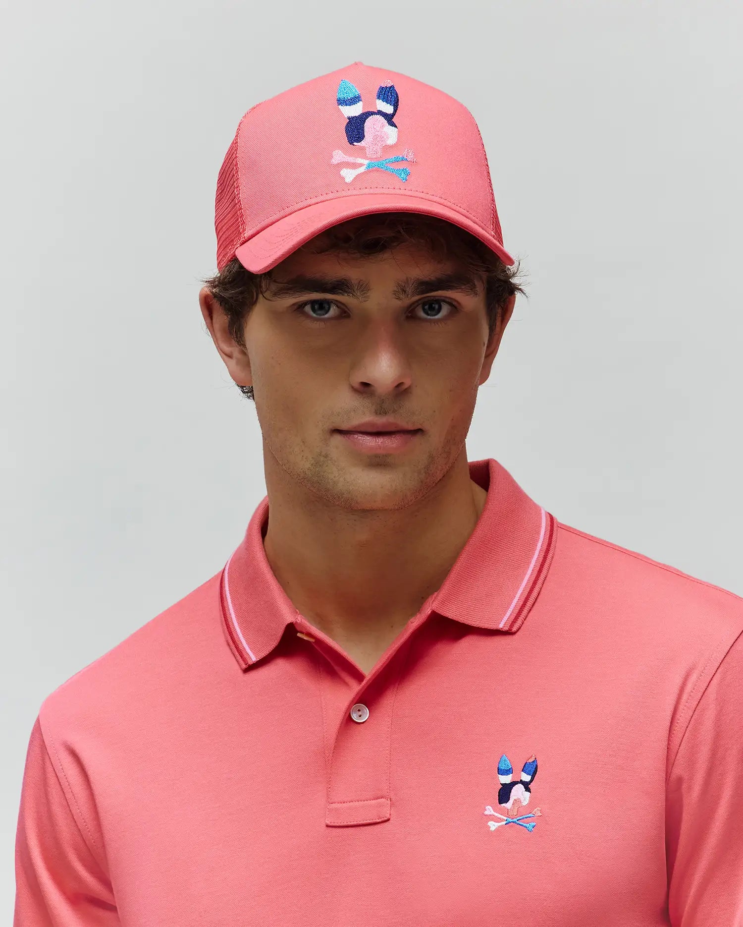 Coral pink pique polo with tipped collar and colorful embroidered Psycho Bunny logo on left chest. Matching Mens Ara Trucker Cap features a large multi-color bunny skull logo. (5)