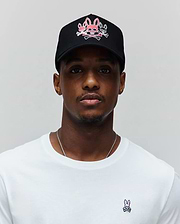 Mens Oscar Trucker Cap in black with a large pink and white embroidered Psycho Bunny skull logo on the front, paired with a white classic fit jersey t-shirt featuring a small embroidered pink and black Psycho Bunny logo on the left chest.