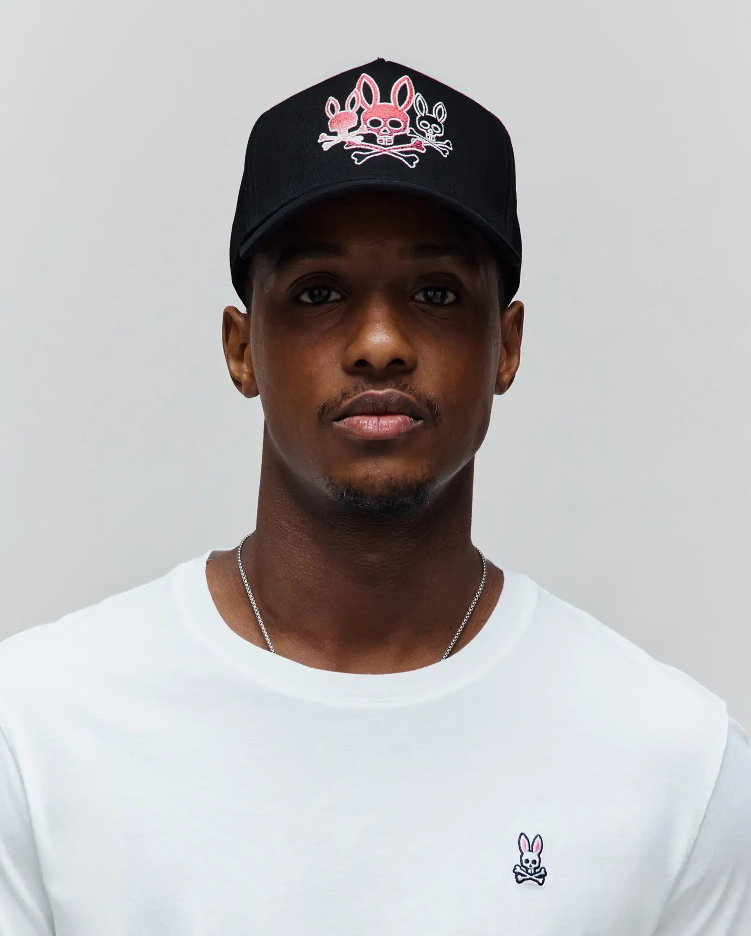 Mens Oscar Trucker Cap in black with a large pink and white embroidered Psycho Bunny skull logo on the front, paired with a white classic fit jersey t-shirt featuring a small embroidered pink and black Psycho Bunny logo on the left chest.