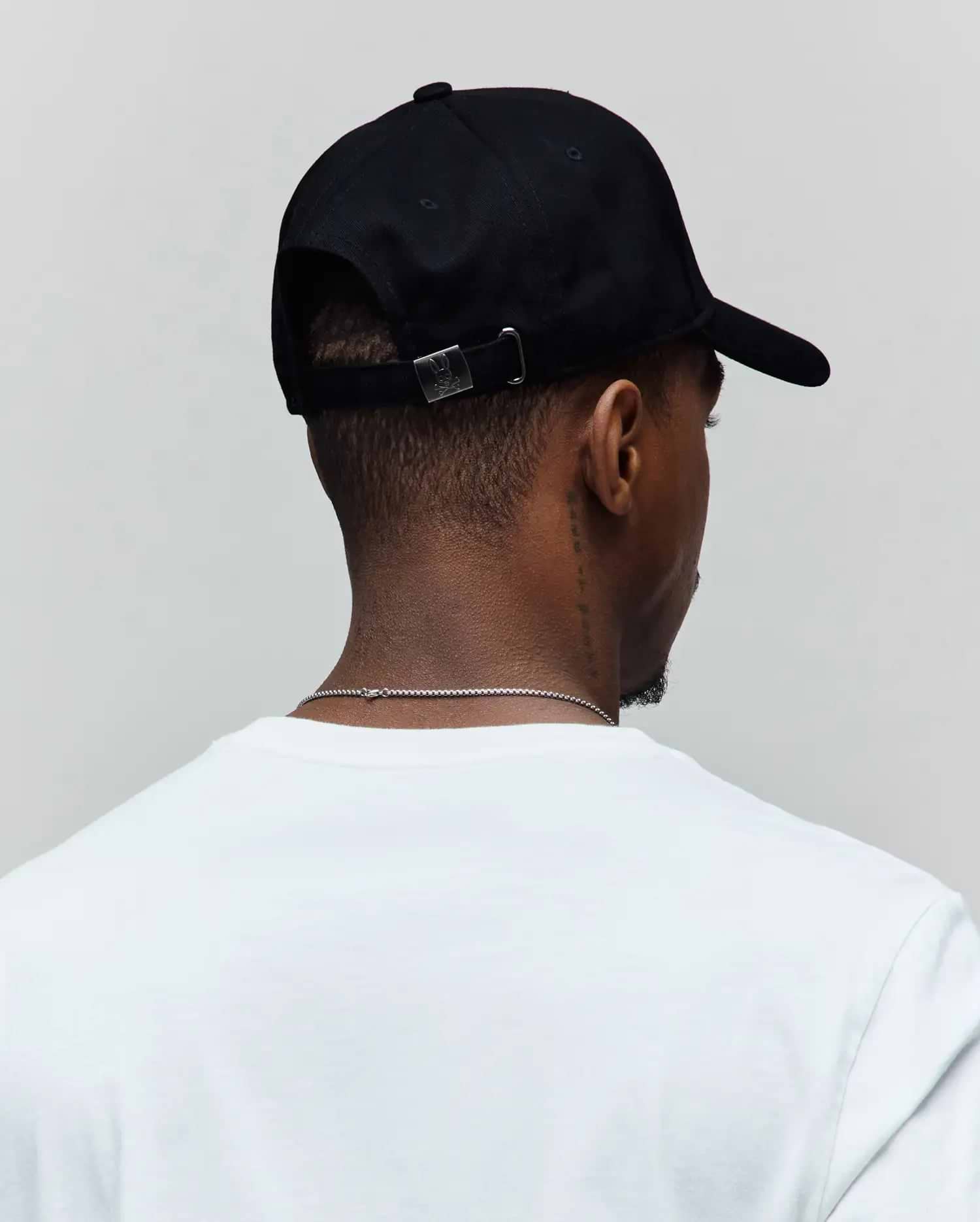 Black Mens Oscar Trucker Cap with a smooth texture, featuring a small silver metal buckle at the back engraved with the iconic Psycho Bunny skull logo, paired with a classic white jersey t-shirt.