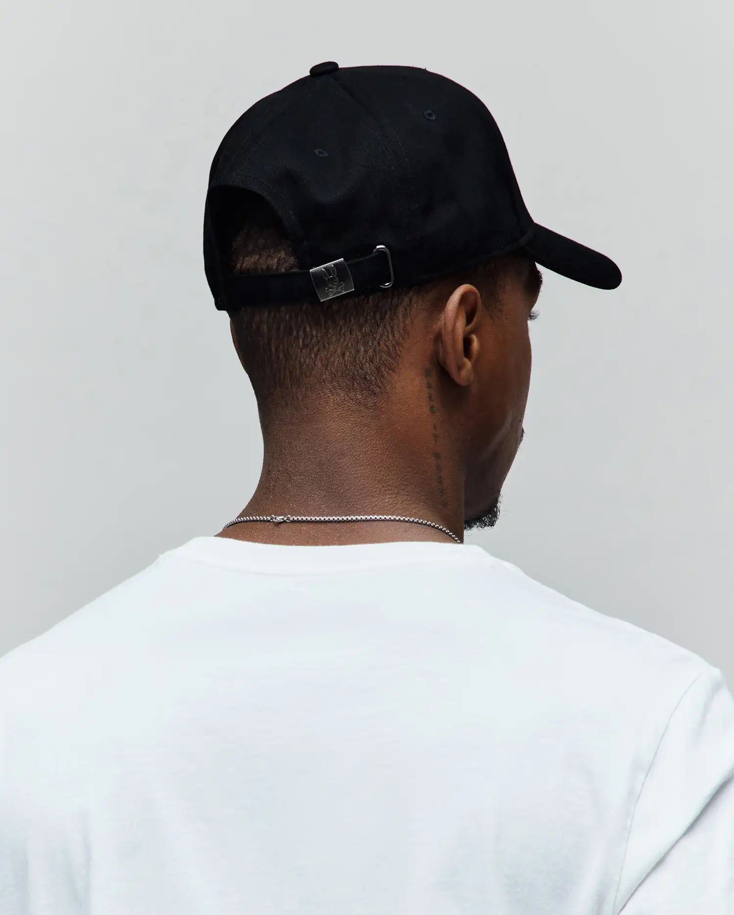 Black Mens Oscar Trucker Cap with a smooth texture, featuring a small silver metal buckle at the back engraved with the iconic Psycho Bunny skull logo, paired with a classic white jersey t-shirt.