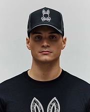 Black Mens Oscar Trucker Cap with white contrast stitching and an embroidered white and pink Psycho Bunny skull logo. Paired with a black jersey t-shirt featuring a large white graphic print. (1)