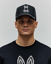 Black Mens Oscar Trucker Cap with white contrast stitching and an embroidered white and pink Psycho Bunny skull logo. Paired with a black jersey t-shirt featuring a large white graphic print. (1)