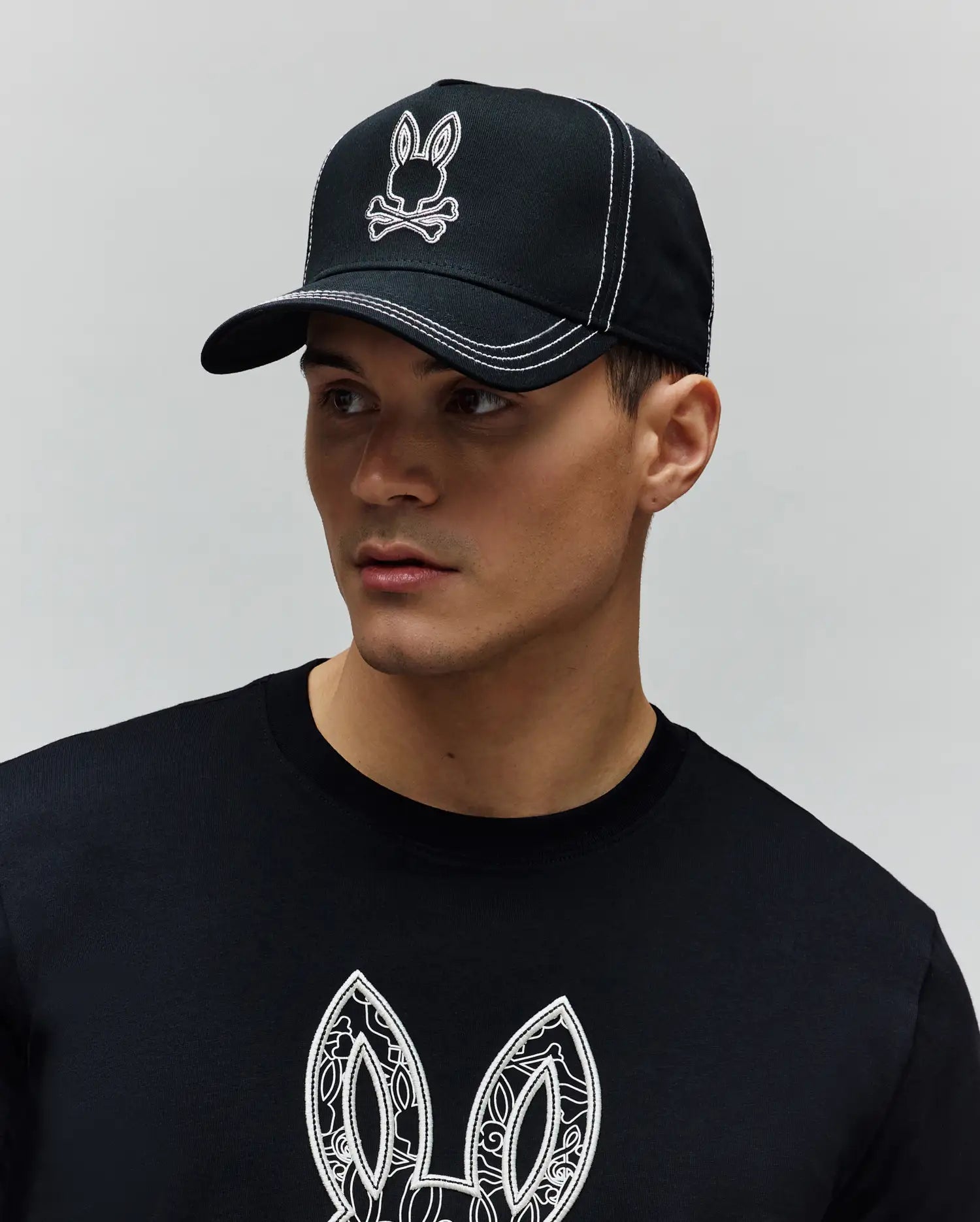 Black Mens Oscar Trucker Cap with white embroidered Psycho Bunny skull logo and white contrast stitching, paired with a black jersey t-shirt featuring a large white patterned Psycho Bunny ear graphic on the chest. (5)