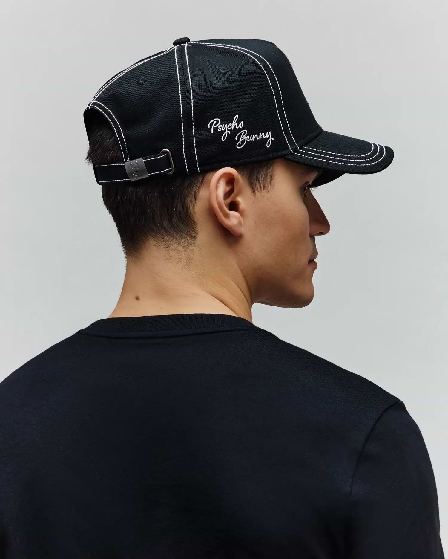 Mens Oscar Trucker Cap, a black cotton twill cap with white contrast stitching, embroidered white Psycho Bunny script logo on the front, and a metal back buckle featuring a bunny skull. A plain black jersey crew neck t-shirt. (3)
