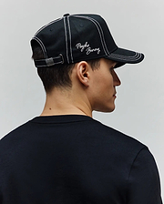Mens Oscar Trucker Cap, a black cotton twill cap with white contrast stitching, embroidered white Psycho Bunny script logo on the front, and a metal back buckle featuring a bunny skull. A plain black jersey crew neck t-shirt. (3)
