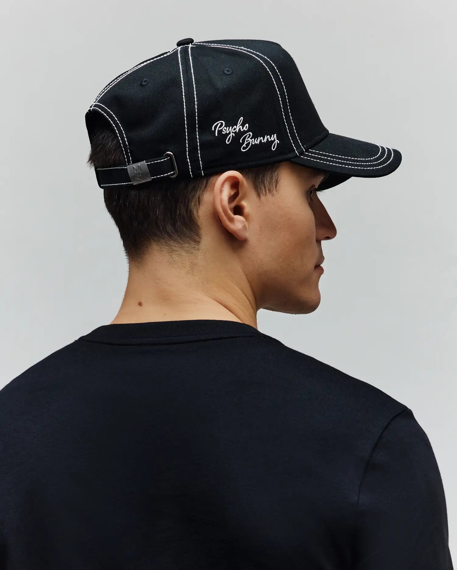 Mens Oscar Trucker Cap, a black cotton twill cap with white contrast stitching, embroidered white Psycho Bunny script logo on the front, and a metal back buckle featuring a bunny skull. A plain black jersey crew neck t-shirt. (3)