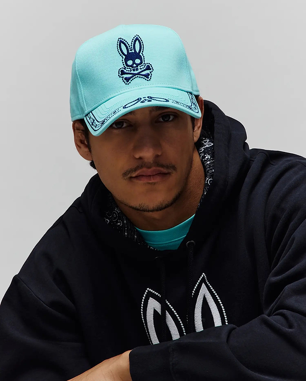 Mens Luke Trucker Cap in aqua with a large dark blue embroidered Psycho Bunny skull logo and bandana brim. Worn over an aqua jersey t-shirt and a black French terry hoodie with white embroidered bunny ears and a bandana-lined hood.