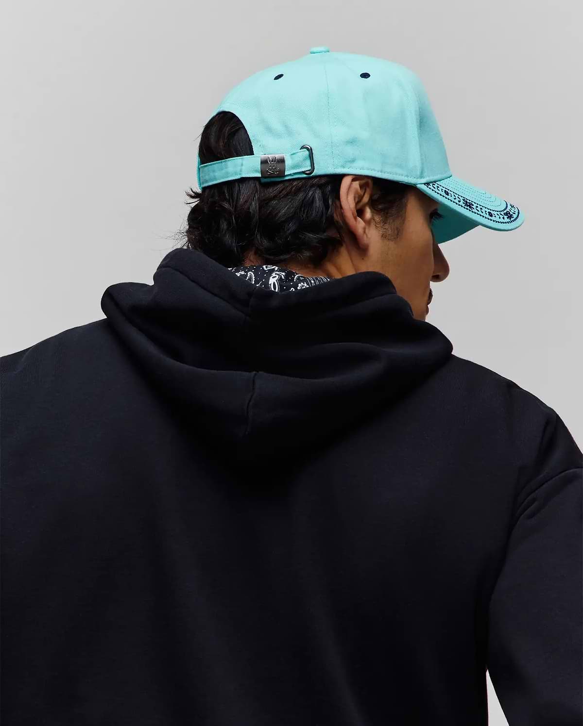 Mens Luke Trucker Cap in aqua with dark blue patterned brim and Psycho Bunny logo buckle, paired with a relaxed fit black french terry hoodie featuring a subtle patterned inner collar. (3)