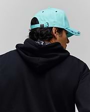 Mens Luke Trucker Cap in aqua with dark blue patterned brim and Psycho Bunny logo buckle, paired with a relaxed fit black french terry hoodie featuring a subtle patterned inner collar. (3)
