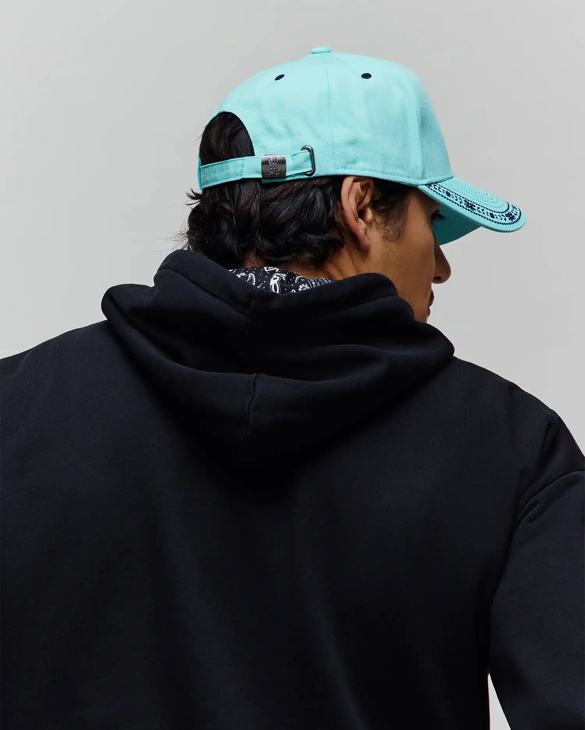 Mens Luke Trucker Cap in aqua with dark blue patterned brim and Psycho Bunny logo buckle, paired with a relaxed fit black french terry hoodie featuring a subtle patterned inner collar. (3)