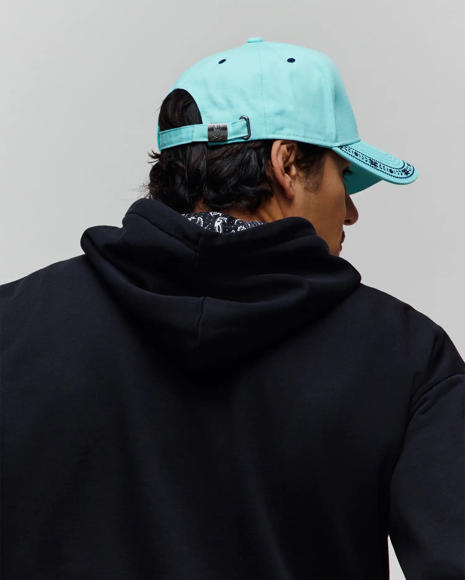 Mens Luke Trucker Cap in aqua with dark blue patterned brim and Psycho Bunny logo buckle, paired with a relaxed fit black french terry hoodie featuring a subtle patterned inner collar. (3)