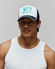 Mens Aram Trucker Cap featuring a white front panel, black mesh back, and a large teal embroidered Psycho Bunny skull logo. The brim is black with a white and teal stripe accent.