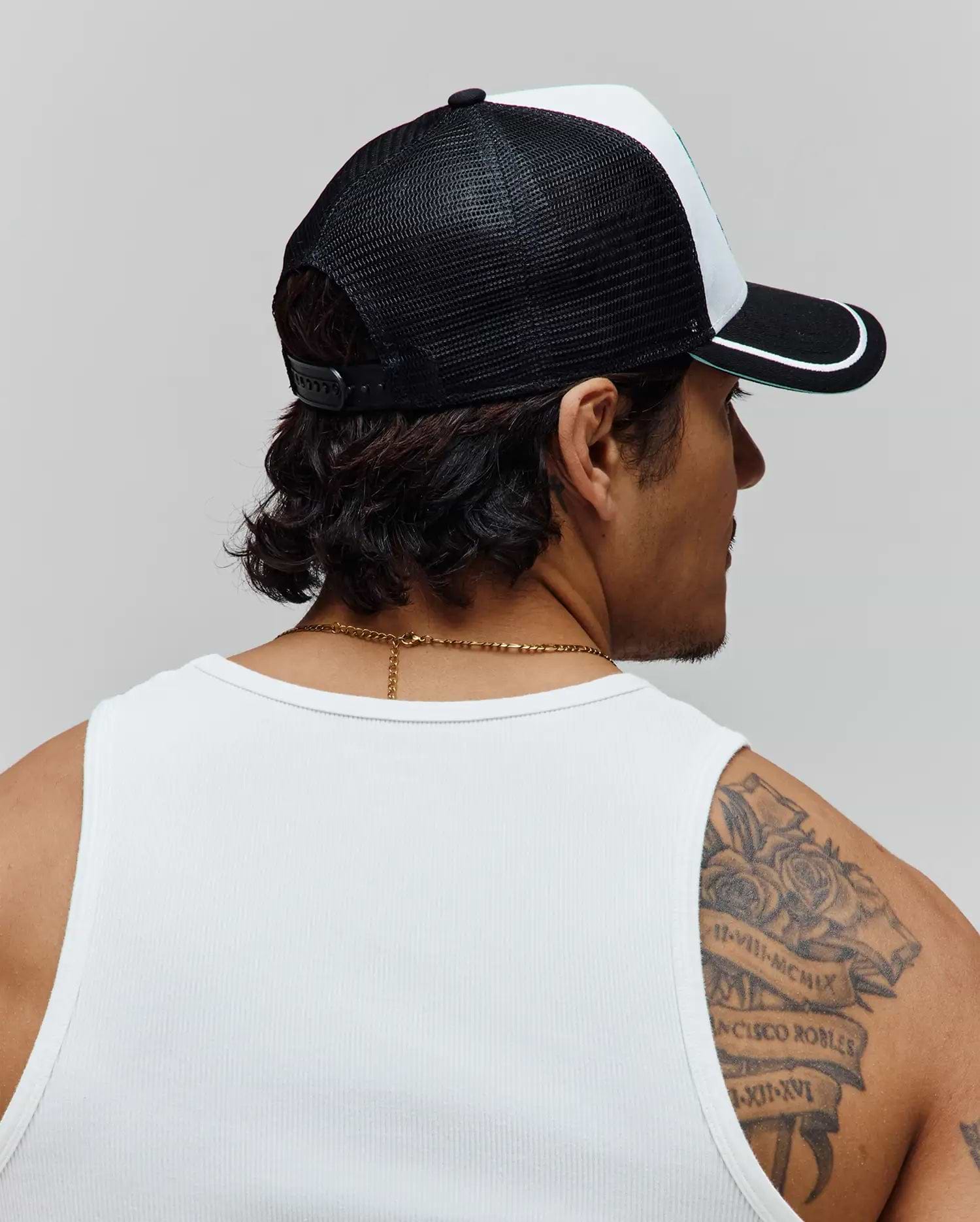 Psycho Bunny Mens Aram Trucker Cap featuring a black mesh back, white front panel, black brim with white stripe and green trim, and an adjustable snapback. Worn with a white ribbed knit tank top.