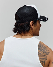 Psycho Bunny Mens Aram Trucker Cap featuring a black mesh back, white front panel, black brim with white stripe and green trim, and an adjustable snapback. Worn with a white ribbed knit tank top.