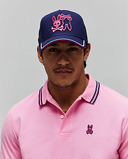 Pink pique polo with navy/pink striped collar and small embroidered Psycho Bunny skull logo on chest. Navy Mens Aram Trucker Cap features large pink embroidered Psycho Bunny logo and white/pink brim stripe. (1)