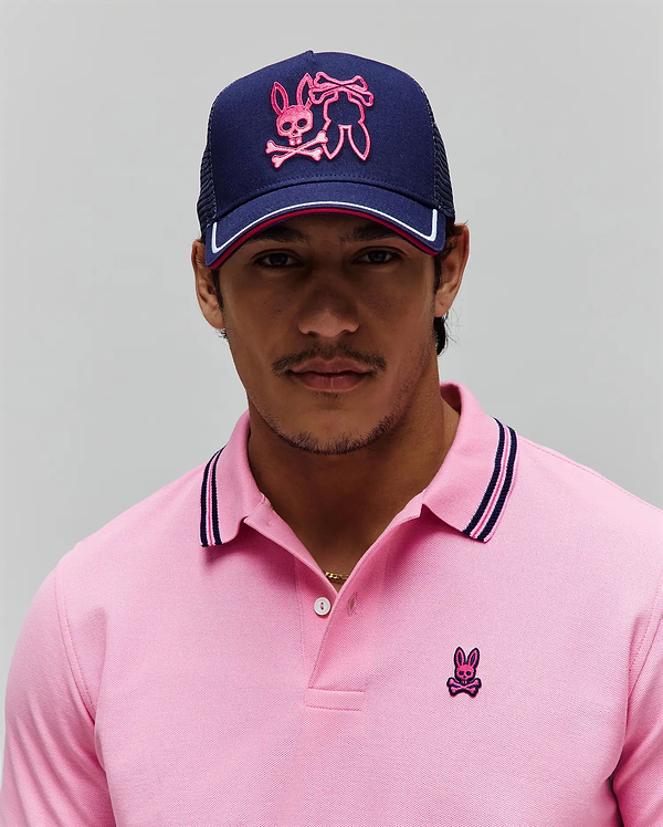 Pink pique polo with navy/pink striped collar and small embroidered Psycho Bunny skull logo on chest. Navy Mens Aram Trucker Cap features large pink embroidered Psycho Bunny logo and white/pink brim stripe.