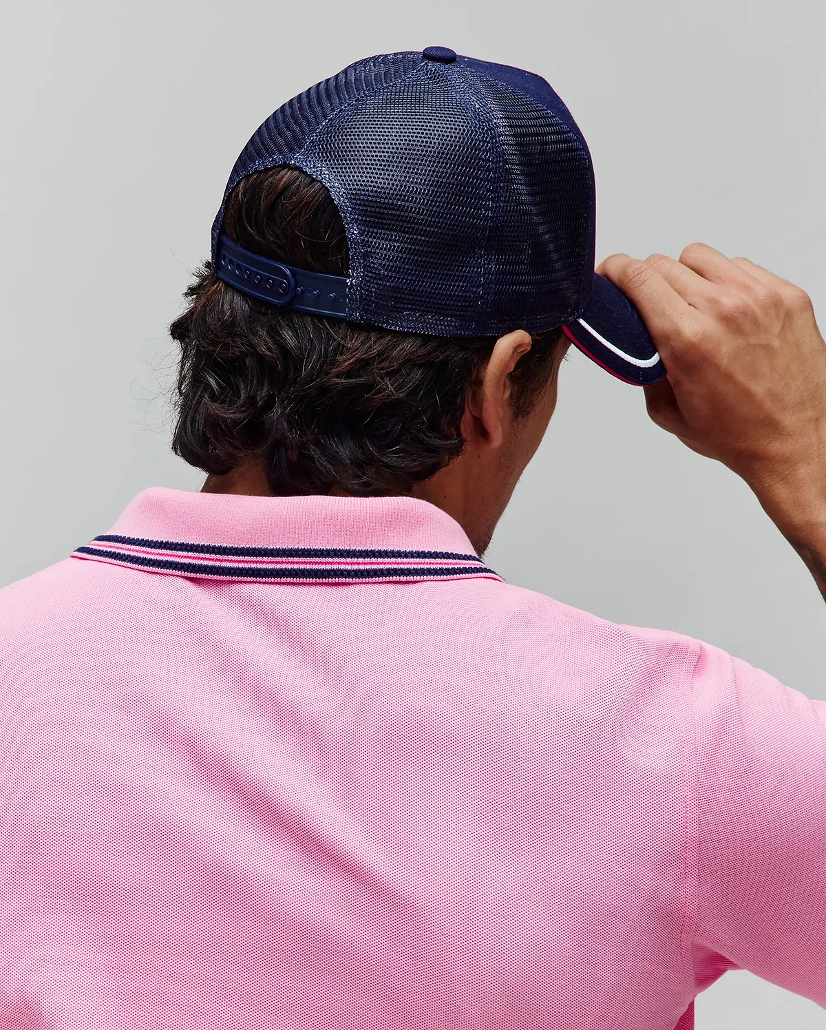 Pink pique polo shirt with navy and pink striped collar, paired with a navy blue mesh Mens Aram Trucker Cap featuring a white and pink striped brim. (3)
