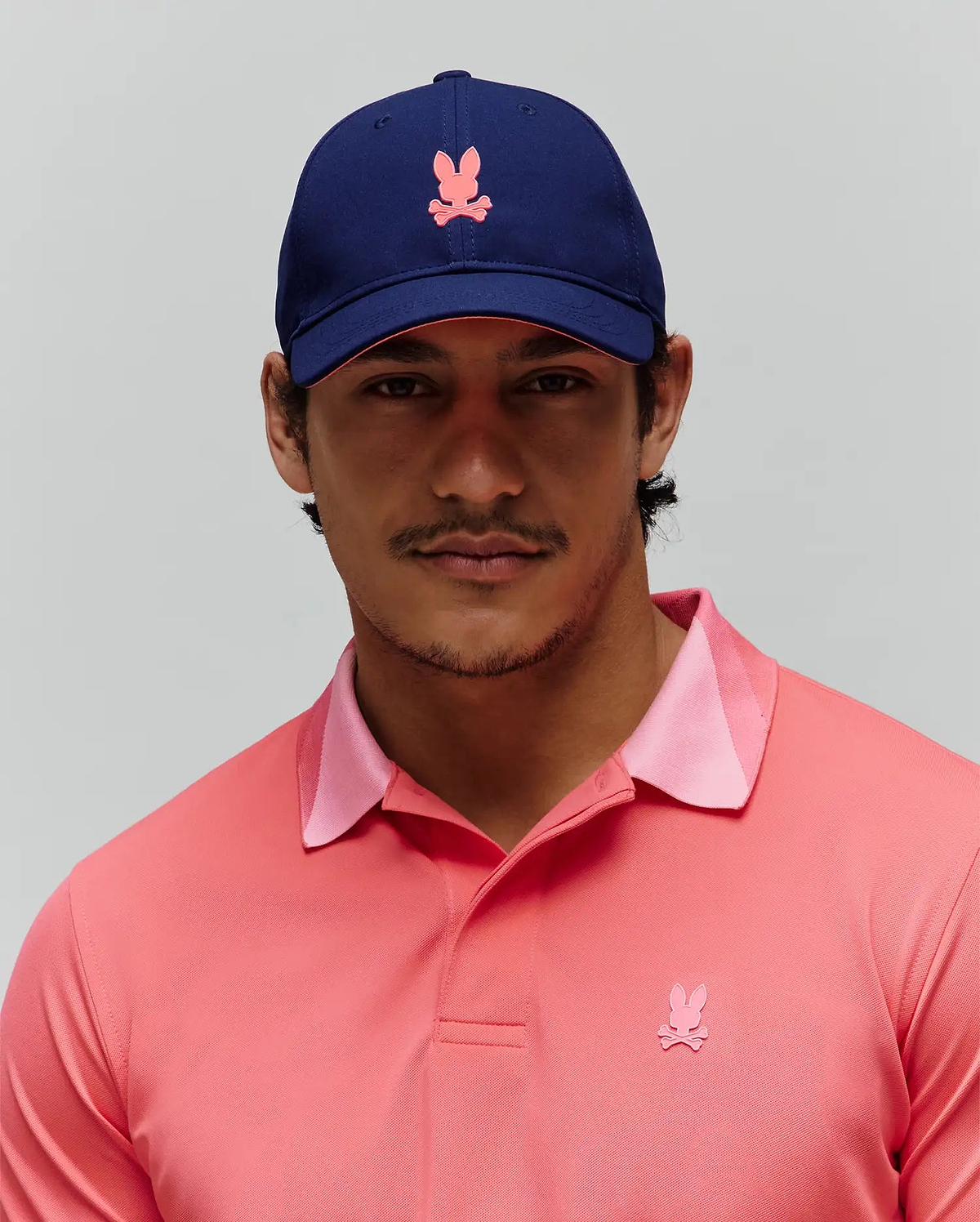 Mens Onyx Sport Cap in navy with a pink embroidered bunny skull logo and a coral pique polo shirt featuring a tonal pink bunny skull logo on the chest and a light pink collar trim. (1)