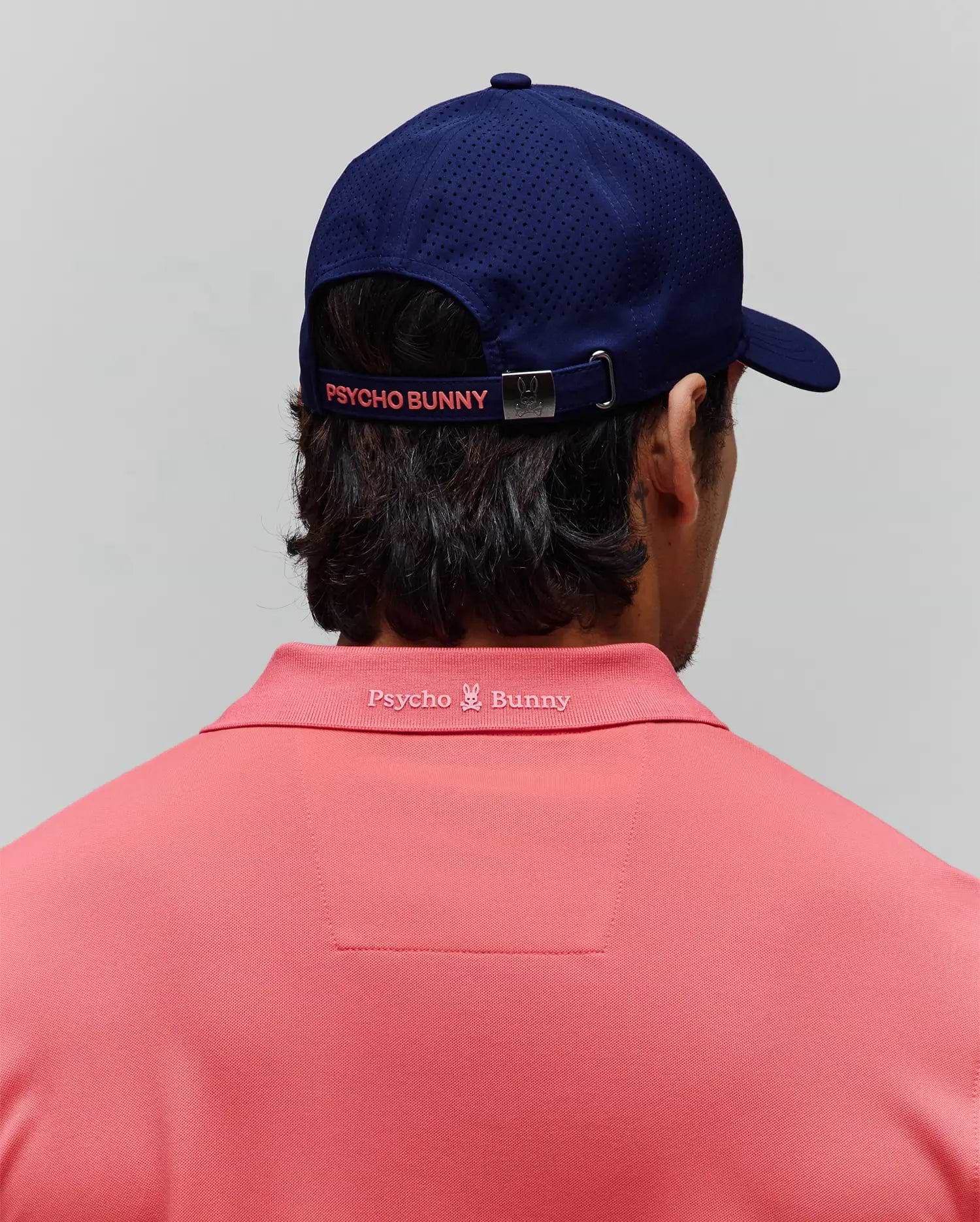 Coral pique polo shirt with tonal embroidered Psycho Bunny logo on the back collar, paired with a navy MENS ONYX SPORT CAP featuring perforated panels and a coral Psycho Bunny logo on the back strap. (3)