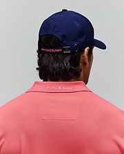 Coral pique polo shirt with tonal embroidered Psycho Bunny logo on the back collar, paired with a navy MENS ONYX SPORT CAP featuring perforated panels and a coral Psycho Bunny logo on the back strap. (3)