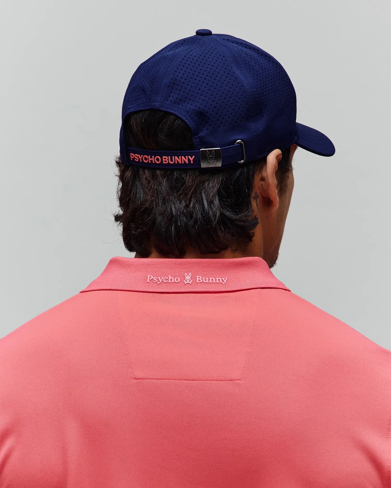 Coral pique polo shirt with tonal embroidered Psycho Bunny logo on the back collar, paired with a navy MENS ONYX SPORT CAP featuring perforated panels and a coral Psycho Bunny logo on the back strap. (3)