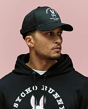 A man stands in front of a pink background, wearing the Psycho Bunny MENS ARCHIVE BASEBALL CAP (MO1500002) and a black 100% cotton hoodie featuring an embroidered bunny logo and "Psycho Bunny" text, looking to the side.