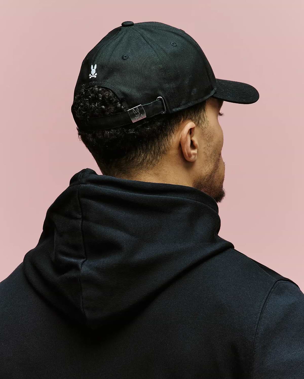 A person wearing a black 100% cotton hoodie and the Psycho Bunny MENS ARCHIVE BASEBALL CAP (MO1500002) is shown from behind against a plain pink background.