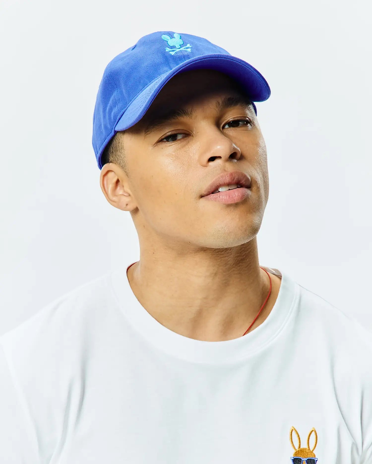 Mens Jimmy Baseball Cap in royal blue with an embroidered light blue Psycho Bunny skull logo, paired with a white jersey t-shirt featuring a small embroidered Psycho Bunny logo wearing sunglasses on the left chest.