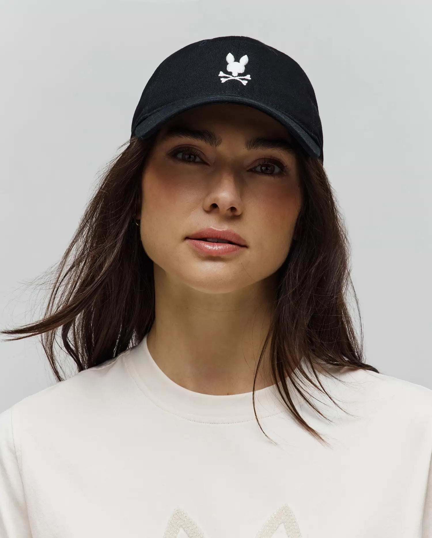 Black Mens Jimmy Baseball Cap with a prominent white embroidered Psycho Bunny skull and crossbones logo on the front, paired with a cream jersey knit t-shirt featuring a subtle textured graphic.