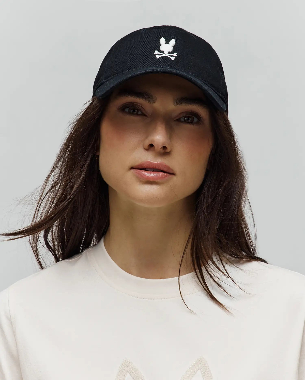 Black Mens Jimmy Baseball Cap with a prominent white embroidered Psycho Bunny skull and crossbones logo on the front, paired with a cream jersey knit t-shirt featuring a subtle textured graphic.
