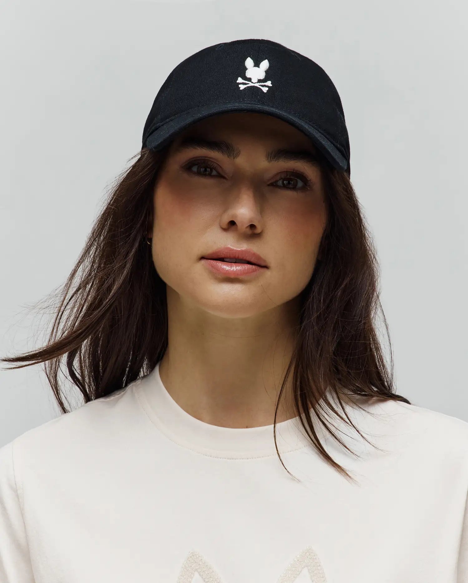 Black Mens Jimmy Baseball Cap with a prominent white embroidered Psycho Bunny skull and crossbones logo on the front, paired with a cream jersey knit t-shirt featuring a subtle textured graphic.