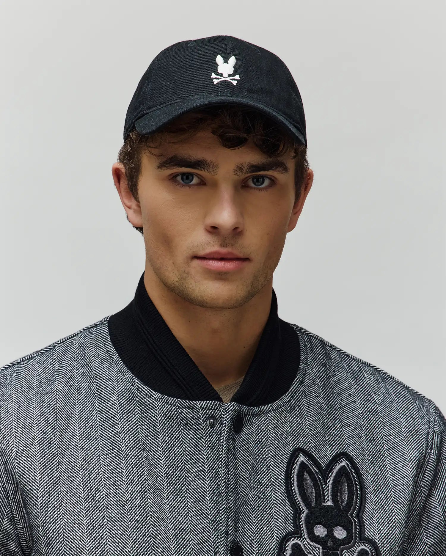Mens Jimmy Baseball Cap in black with a white embroidered Psycho Bunny skull logo, paired with a black and white herringbone bomber jacket featuring a large embroidered Psycho Bunny skull patch.