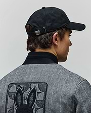Black Mens Jimmy Baseball Cap with a metal Psycho Bunny buckle, paired with a grey herringbone jacket featuring a black ribbed collar and a large embroidered Psycho Bunny skull logo on the back.