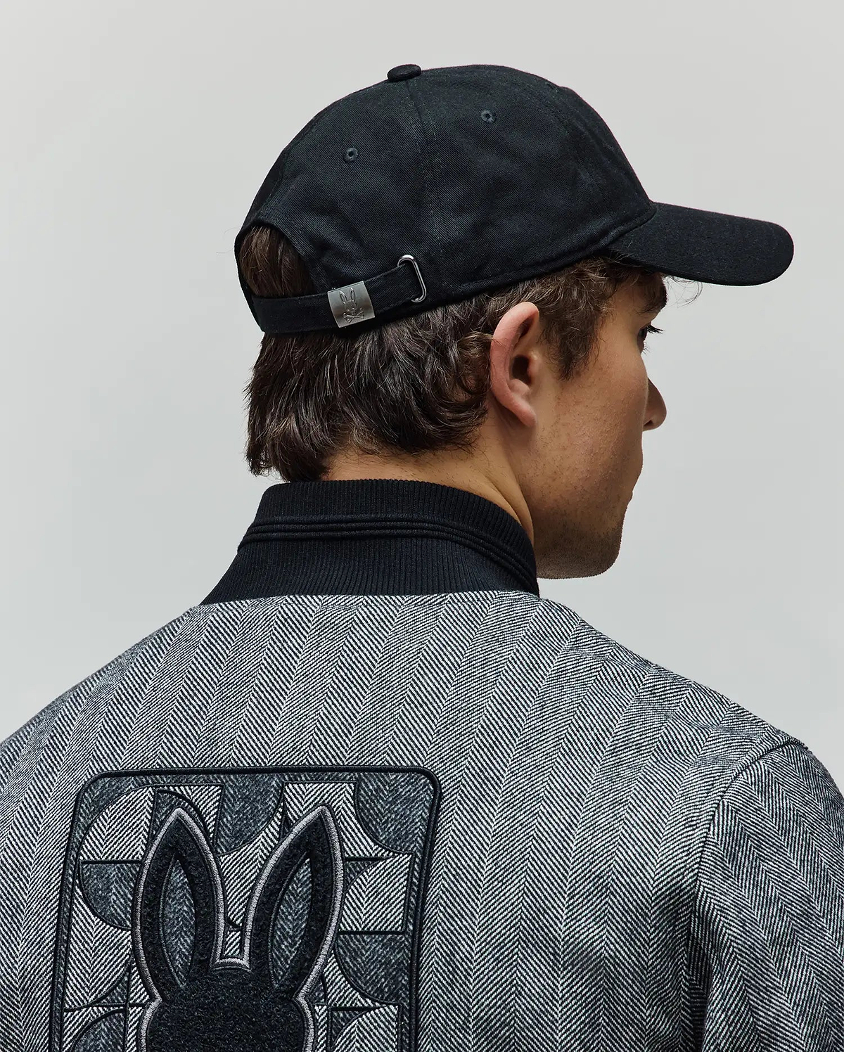 Black Mens Jimmy Baseball Cap with a metal Psycho Bunny buckle, paired with a grey herringbone jacket featuring a black ribbed collar and a large embroidered Psycho Bunny skull logo on the back.