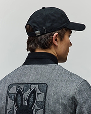 Black Mens Jimmy Baseball Cap with a metal Psycho Bunny buckle, paired with a grey herringbone jacket featuring a black ribbed collar and a large embroidered Psycho Bunny skull logo on the back.
