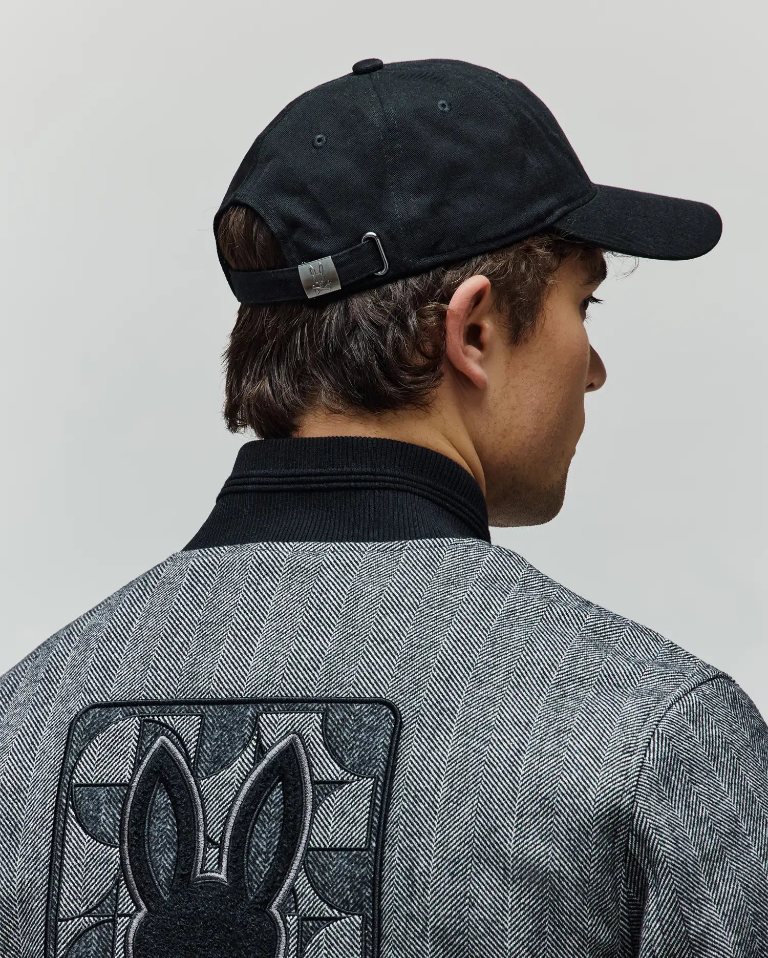 Black Mens Jimmy Baseball Cap with a metal Psycho Bunny buckle, paired with a grey herringbone jacket featuring a black ribbed collar and a large embroidered Psycho Bunny skull logo on the back.