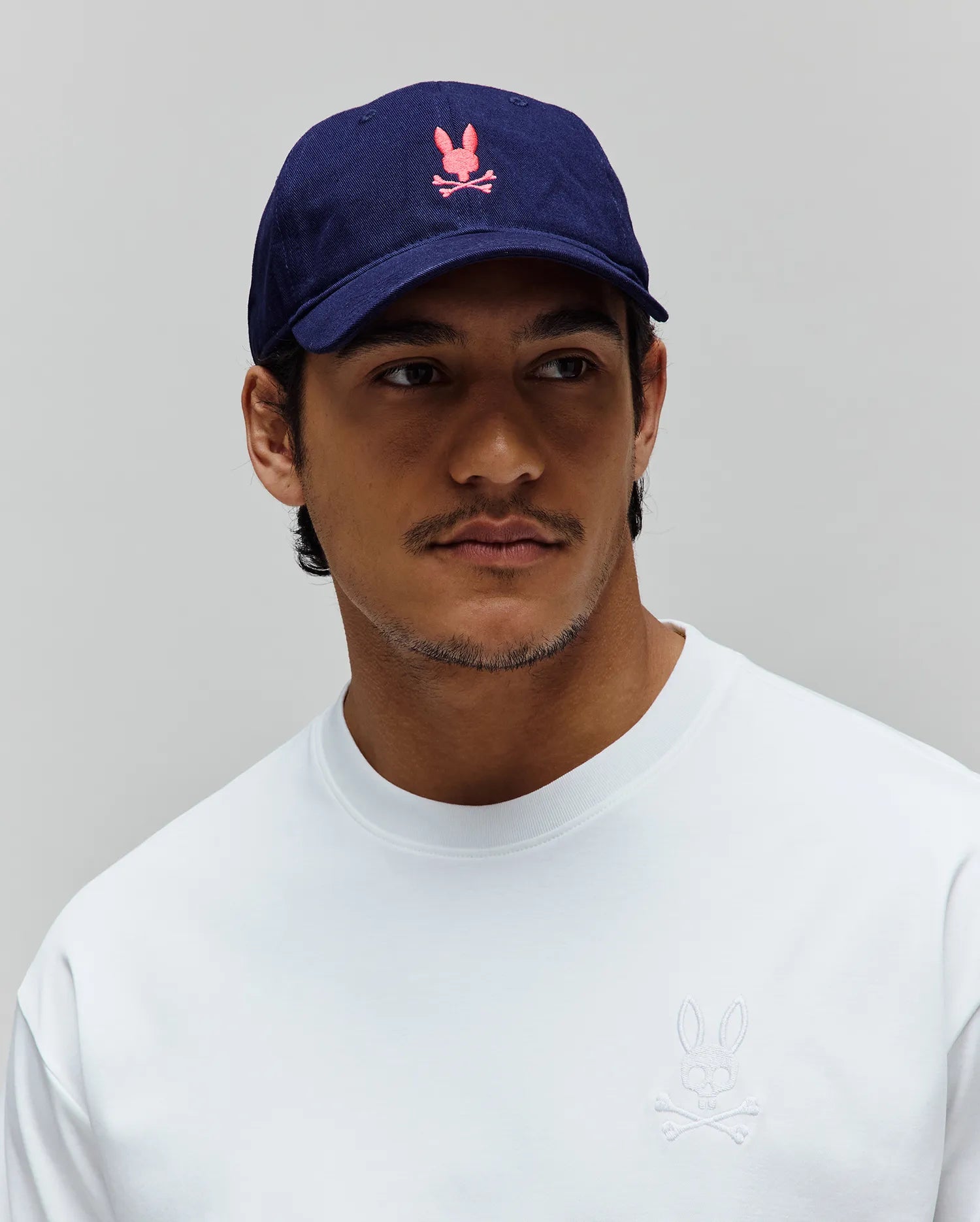 Navy cotton twill Mens Jimmy Baseball Cap with a pink embroidered Psycho Bunny skull logo, and a white relaxed-fit jersey crewneck t-shirt with a small white embroidered Psycho Bunny logo on the left chest.