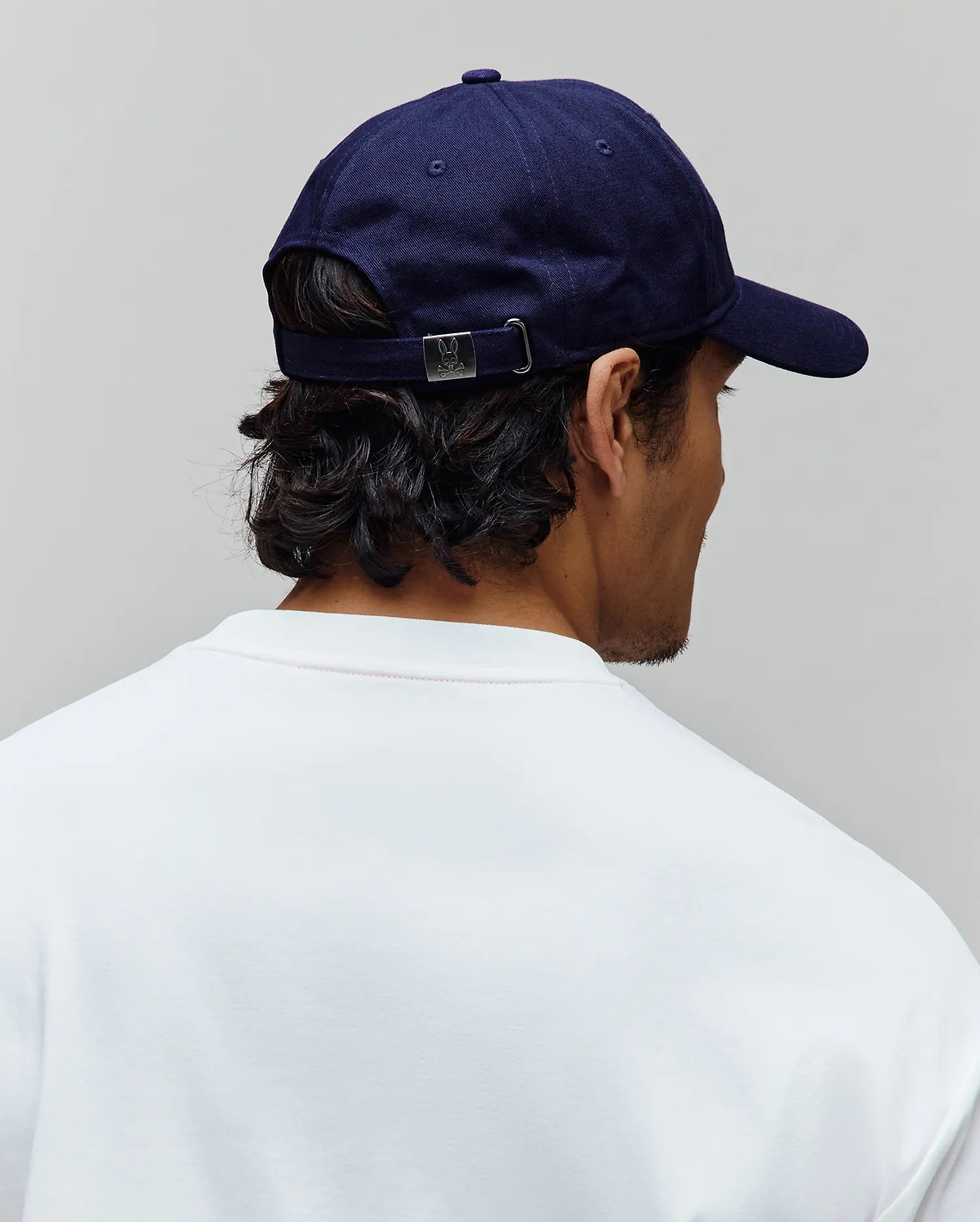 Mens Jimmy Baseball Cap in navy blue cotton twill, featuring a silver metal buckle with the iconic Psycho Bunny skull logo, worn with a crisp white jersey t-shirt.