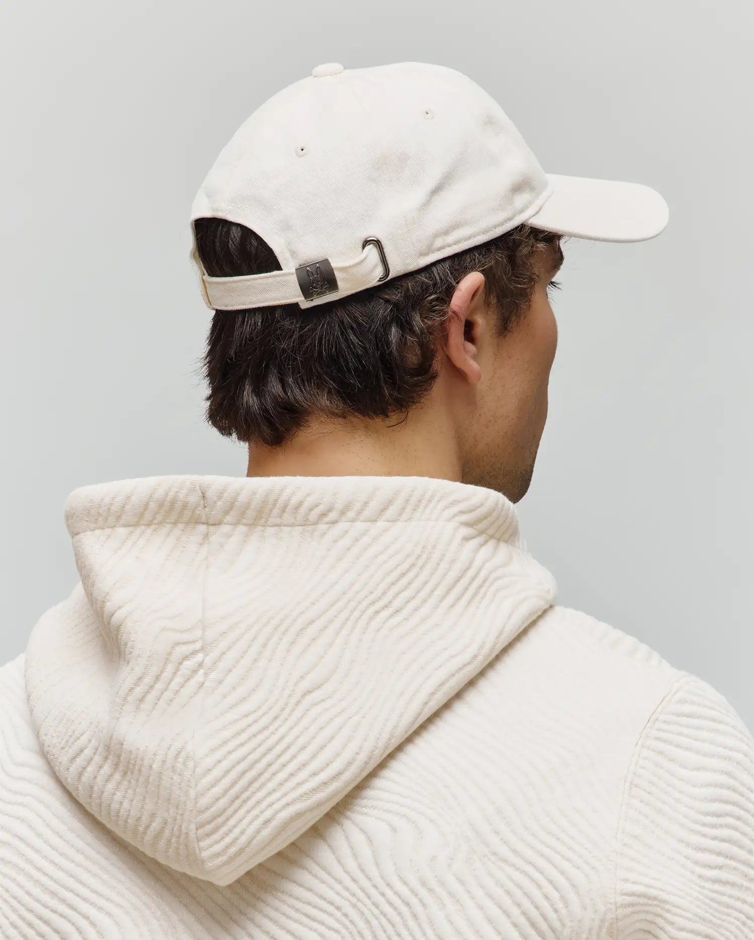 Mens Jimmy Baseball Cap in cream cotton twill with a dark metal buckle featuring a small Psycho Bunny skull logo, paired with a cream hoodie in a unique wavy textured knit.