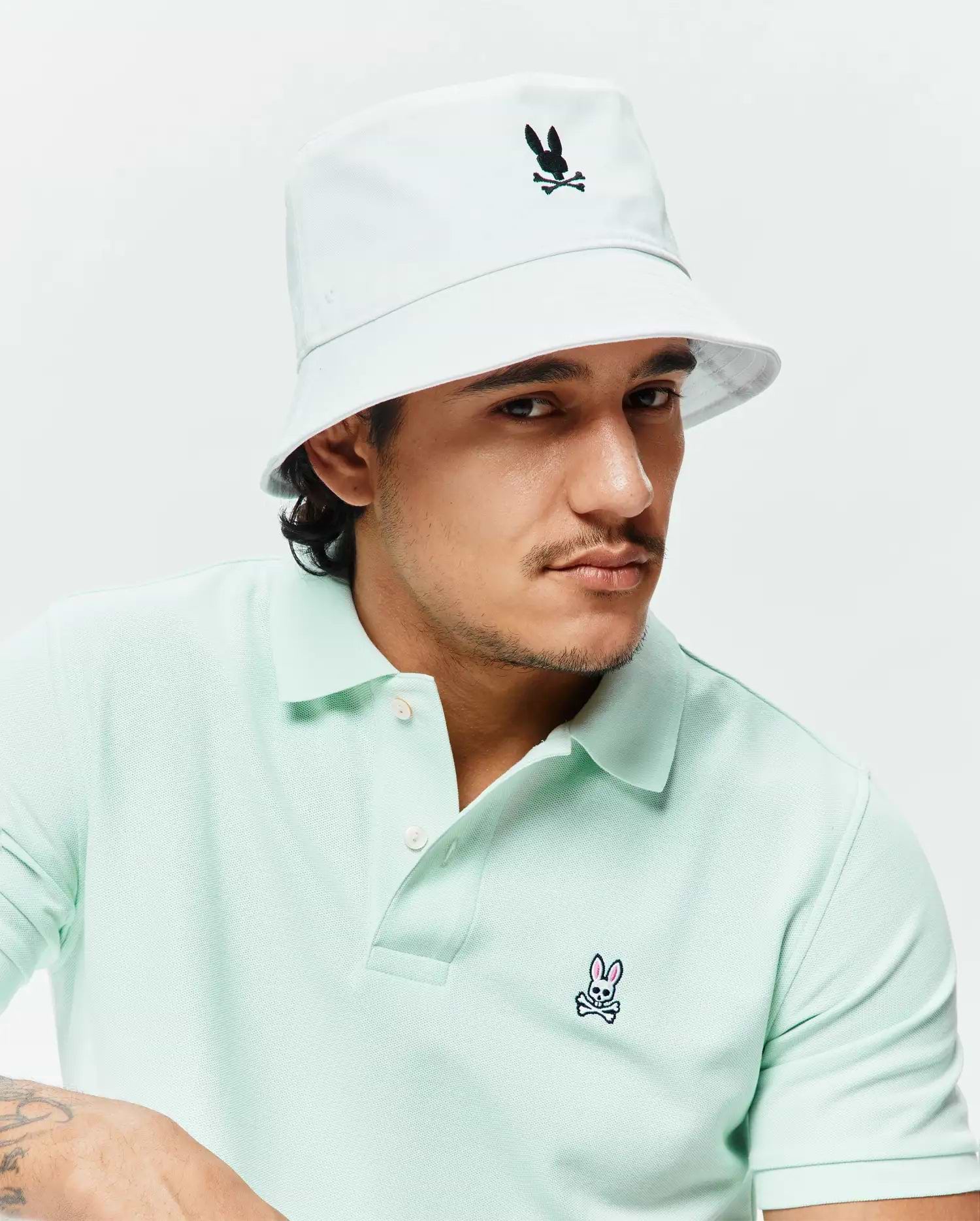 Light mint green pique polo shirt with a small embroidered Psycho Bunny logo on the chest, paired with a white Mens Jimmy Bucket Hat featuring a black embroidered bunny skull.