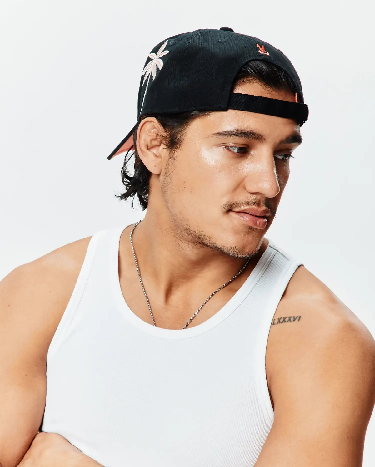 Black Mens Brad Trucker Cap featuring a front light pink embroidered palm tree, small orange Psycho Bunny logo on the back, and peach brim underside, alongside a white ribbed knit classic fit tank top.