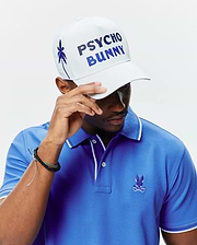 Royal blue pique polo shirt with white tipping on collar and cuffs, featuring a small embroidered Psycho Bunny skull logo on the chest. Paired with a white MENS BRAD TRUCKER CAP with blue gradient PSYCHO BUNNY text and an embroidered palm tree.
