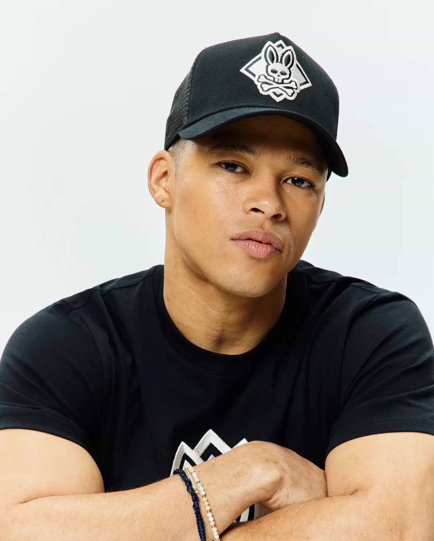 Black Mens Tobias Trucker Cap with a white embroidered Psycho Bunny skull and crossbones logo on the front, featuring a mesh back. Worn with a black classic fit jersey t-shirt displaying a large white geometric chest graphic.