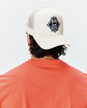 Mens Tobias Trucker Cap in cream with a prominent navy and black embroidered Psycho Bunny skull logo within a diamond on the front, featuring a mesh back. Paired with a coral jersey knit t-shirt.