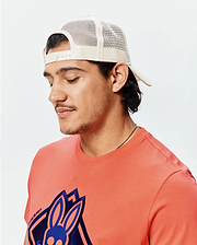 Coral jersey t-shirt with a large, layered dark and light blue embroidered Psycho Bunny skull logo and vertical text on the chest, paired with a cream mesh Mens Tobias Trucker Cap.
