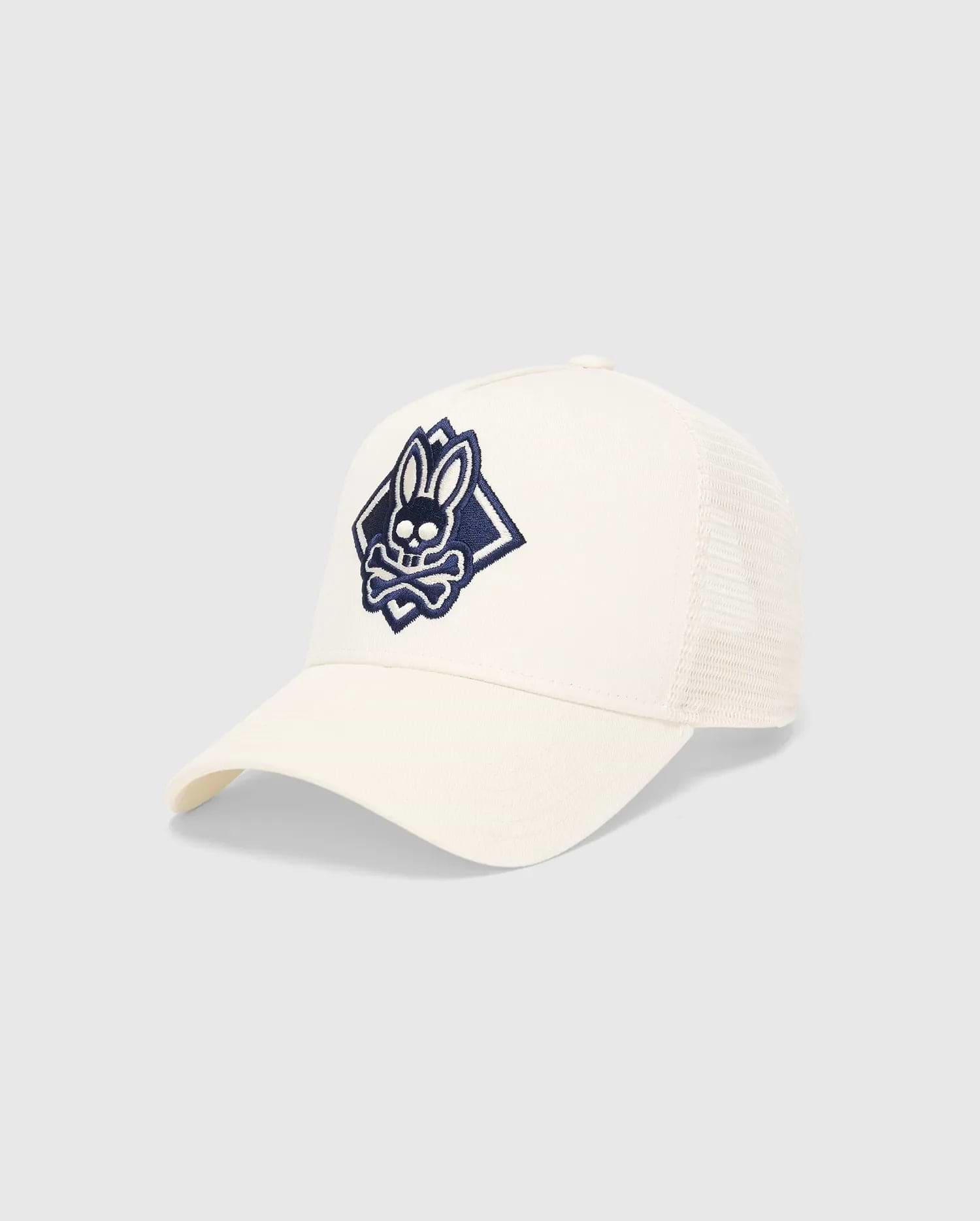 Mens Tobias Trucker Cap in cream, featuring a large, embroidered navy blue Psycho Bunny skull logo within a diamond on the front, with a cotton twill front and breathable mesh back panels.