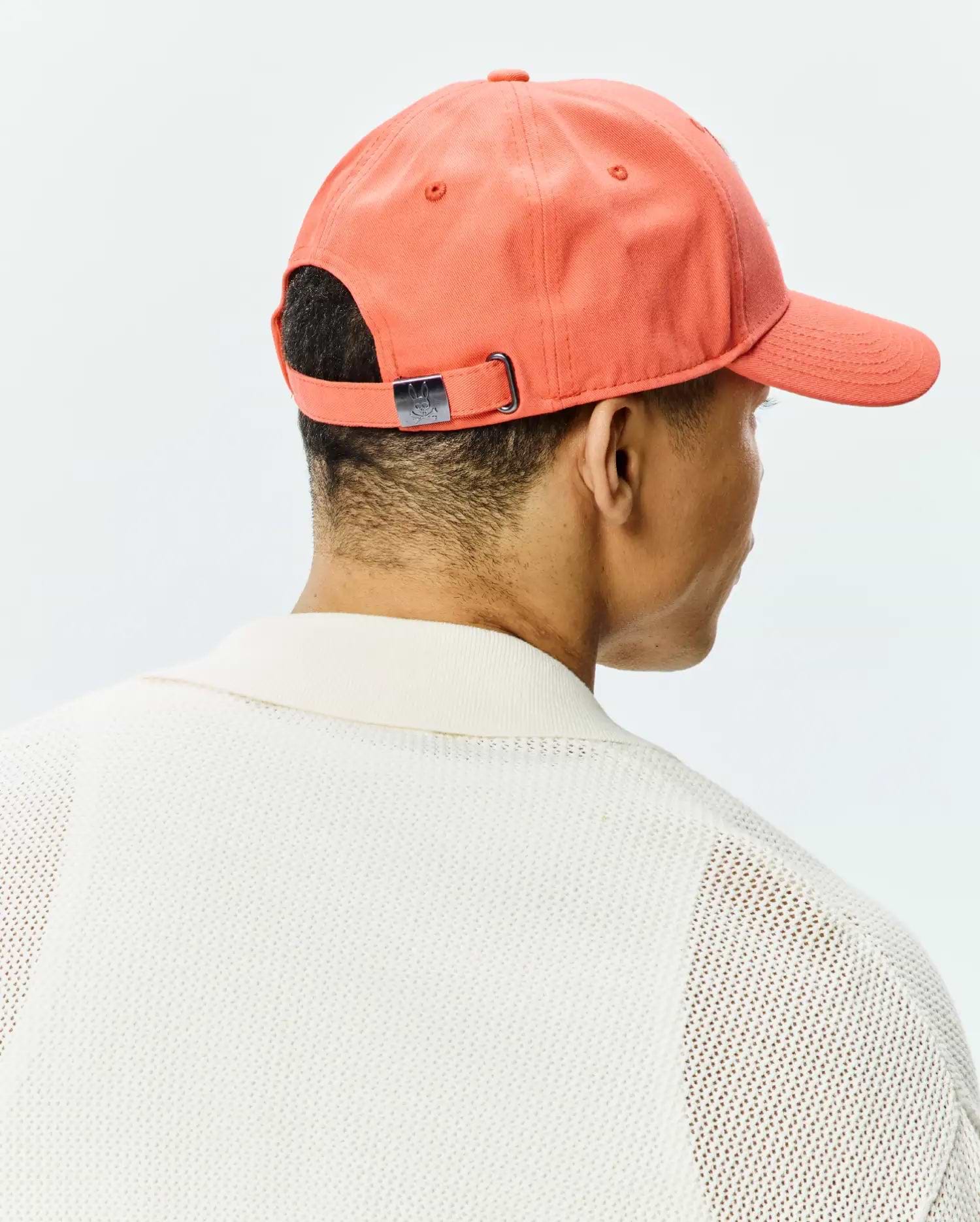 Vibrant coral Mens Craig Baseball Cap in cotton twill with a metallic Psycho Bunny skull logo buckle, worn with a cream open-knit polo sweater featuring a ribbed collar.