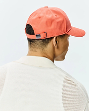 Vibrant coral Mens Craig Baseball Cap in cotton twill with a metallic Psycho Bunny skull logo buckle, worn with a cream open-knit polo sweater featuring a ribbed collar.
