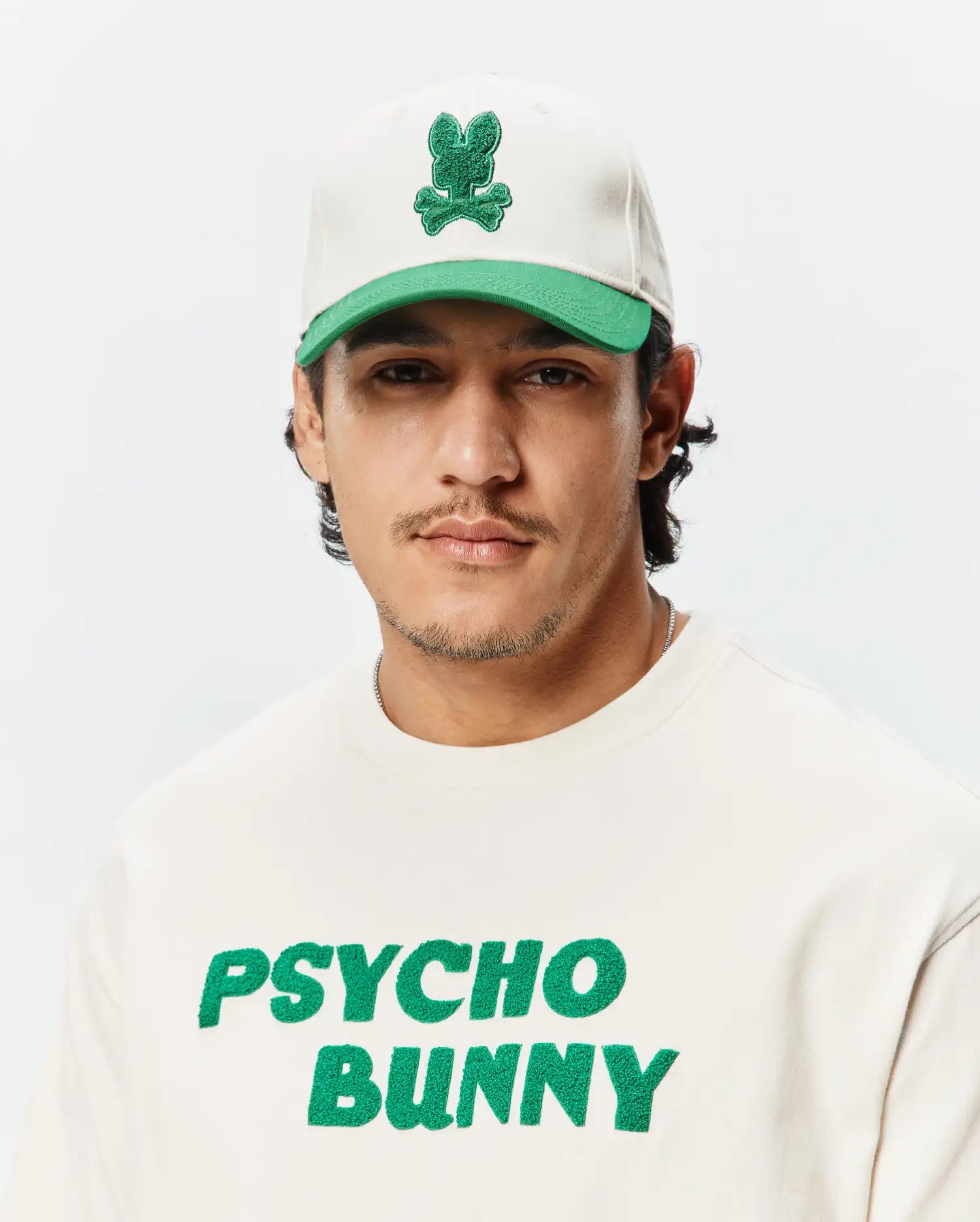 Mens Craig Baseball Cap in off-white with a green brim and embroidered green bunny skull logo, paired with an off-white crewneck sweatshirt featuring a large green chenille Psycho Bunny text logo.